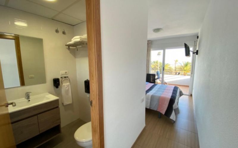 Vista del interior de una de las habitaciones con el baño en primer plano, la cama con televisor y puerta de terraza al fondo